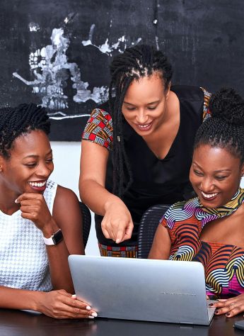 Three African American women collaborating at a laptop, focused and happy during a meeting indoors.